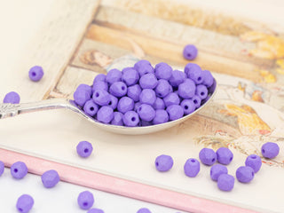 A silver spoon holds saturated, opaque, purple Czech glass beads for jewellery, 4mm matte faceted spacers (50 pcs), with more fire-polished beads scattered on a pastel-illustrated book — ideal for tasselled earrings.