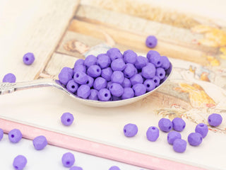 A silver spoon holds saturated, opaque, purple Czech glass beads for jewellery, 4mm matte faceted spacers (50 pcs), with more fire-polished beads scattered on a pastel-illustrated book — ideal for tasselled earrings.