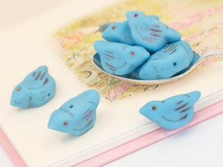 A silver spoon holds several large pressed Czech glass songbird beads in opaque matte sky blue with metallic rainbow volcano wash (11x22mm), resting on a pastel illustrated book page. A few more bird bead DIY earring components are placed nearby.